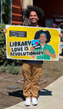 Mychal Threets, a Black librarian, is pictured holding a giant library card from Berkeley Public Library. The card is yellow, and has the words LIBRARY LOVE IS REVOLUTIONARY on it. The image is of a cartoon Mychal holding a book with the title I Love My Hair! 