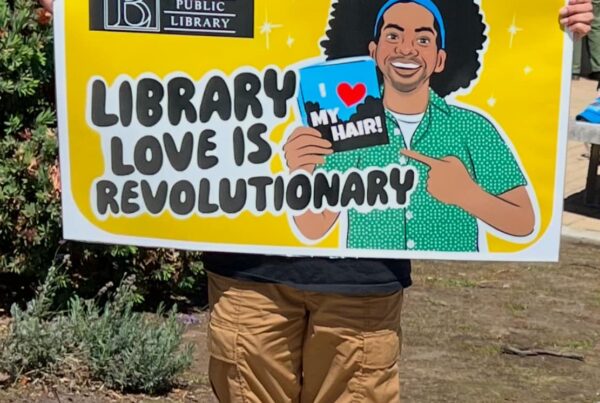 Mychal Threets, a Black librarian, is pictured holding a giant library card from Berkeley Public Library. The card is yellow, and has the words LIBRARY LOVE IS REVOLUTIONARY on it. The image is of a cartoon Mychal holding a book with the title I Love My Hair!